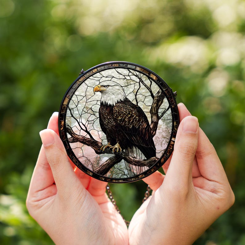 May include: A circular stained glass window depicting a bald eagle on a tree branch. The eagle has a white head and brown body with a yellow beak. The window is framed in black and has a chain for hanging.