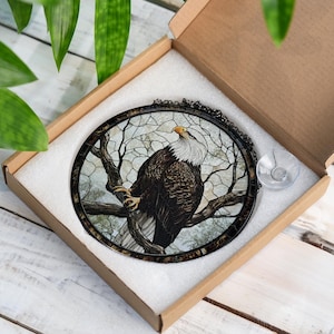 May include: Circular stained glass artwork depicting a bald eagle on a tree branch. The eagle has a white head and yellow beak, with brown and white feathers. The artwork is set in a black metal frame and comes in a cardboard box.