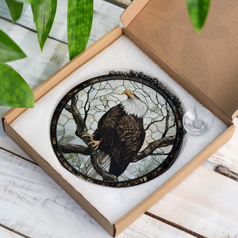 May include: Circular stained glass artwork depicting a bald eagle on a tree branch. The eagle has a white head and yellow beak, with brown and white feathers. The artwork is set in a black metal frame and comes in a cardboard box.