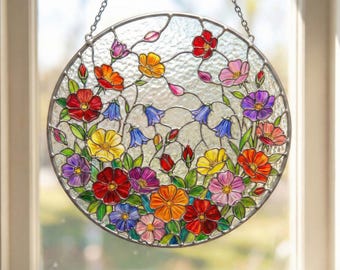 May include: A round stained glass window hanging with a floral design. The window features a variety of colorful flowers in shades of red, orange, yellow, pink, and purple, with green leaves and blue bell-shaped flowers. The window is framed with a silver metal border and hangs from a chain.