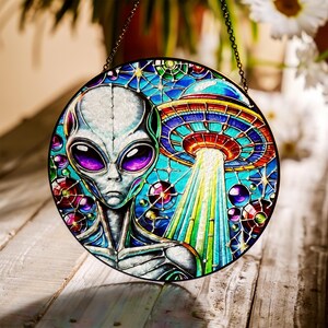 May include: Round stained glass window featuring an alien with large purple eyes and a UFO. The artwork has a colorful design with blue, green, red, and purple hues. The window is hanging from a chain.