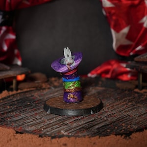May include: A white rabbit figurine peeking out of a purple, green, and gold hat, sitting on a black circular base. The figurine is on a wooden stage with red fabric curtains in the background.