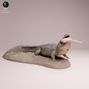 Pode incluir: Escultura detalhada de um jacaré com um tubarão na boca, repousando sobre uma base arenosa. O jacaré é cinza com pele texturizada, e o tubarão é branco. A escultura é uma miniatura, provavelmente para exibição ou coleção.