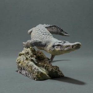Pode incluir: Uma escultura detalhada de um jacaré em uma pose dinâmica, empoleirada em uma formação rochosa texturizada. O jacaré é principalmente cinza com uma pele texturizada, e a rocha tem uma mistura de tons verdes e marrons. O fundo é cinza liso.