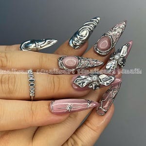 May include: A hand displaying long, pointed, pink and silver nail art. The nails feature intricate silver designs, including swirls, floral accents, and geometric patterns. A silver ring with small, connected stones is also visible. The text "14nailsart" is present.