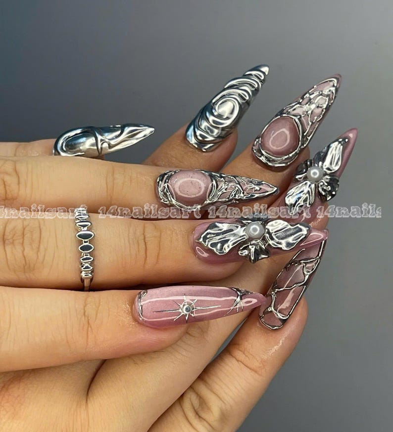 May include: A hand displaying long, pointed, pink and silver nail art. The nails feature intricate silver designs, including swirls, floral accents, and geometric patterns. A silver ring with small, connected stones is also visible. The text "14nailsart" is present.