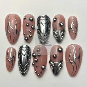 May include: A set of ten artificial nails in various designs. Some nails are a nude pink with silver accents, while others feature silver embellishments, including spheres and heart-shaped designs. The nails are oval-shaped.