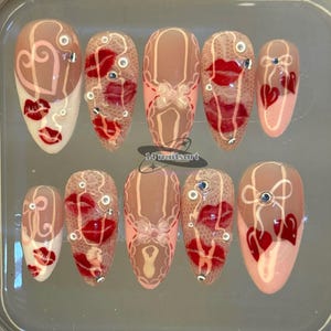 Pode incluir: Um conjunto de dez unhas artificiais com v&aacute;rios designs. As unhas apresentam uma base nude com detalhes de l&aacute;bios vermelhos, cora&ccedil;&otilde;es e padr&otilde;es de renda. Algumas unhas t&ecirc;m pequenos strass decorativos. O texto "14 nailsart" &eacute; vis&iacute;vel.