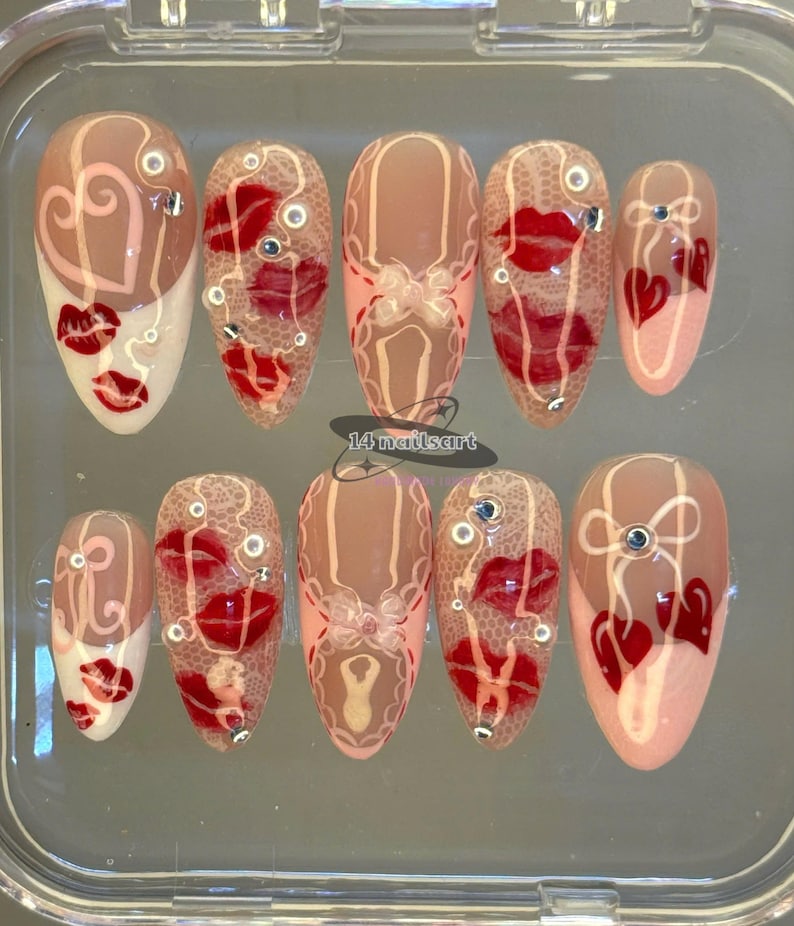 Pode incluir: Um conjunto de dez unhas artificiais com v&aacute;rios designs. As unhas apresentam uma base nude com detalhes de l&aacute;bios vermelhos, cora&ccedil;&otilde;es e padr&otilde;es de renda. Algumas unhas t&ecirc;m pequenos strass decorativos. O texto "14 nailsart" &eacute; vis&iacute;vel.