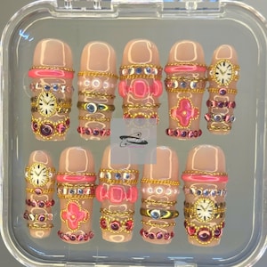 Puede incluir: Un conjunto de uñas artificiales en una funda transparente. Las uñas son de color nude con varios adornos, incluyendo cadenas doradas, detalles rosas y detalles de joyas. Algunas uñas presentan pequeñas esferas de reloj.