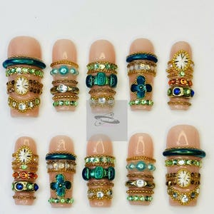 May include: Ten artificial nails with a nude base, each decorated with elaborate, jewel-toned adornments. Designs feature gold chains, emerald green details, and tiny clock faces, creating a luxurious and ornate appearance.