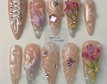 May include: A set of ten almond-shaped, artificial nails. Each nail features a unique design, including lace-up details, floral accents, and sparkling embellishments. The nails are primarily a soft pink with silver and white accents.