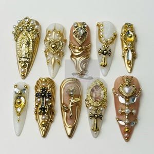 Può includere: Una collezione di dieci unghie finte lunghe e appuntite. Ogni unghia presenta disegni intricati con oro, perle e strass. I disegni includono croci, cuori e figure religiose, creando un'estetica lussuosa e ornata.