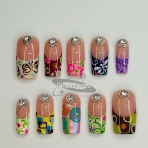 May include: A set of ten artificial nails with various colorful designs. Each nail features a clear, rounded tip and a decorative pattern, including butterflies, flowers, and geometric shapes. A small, silver gem is affixed to the base of each nail. The text "14nailsart" is visible.