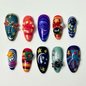 May include: A collection of ten artificial nails with various designs. The nails feature a variety of colors, including blue, red, orange, and purple. Some nails have 3D embellishments like a dragonfly, stars, and a spider. The nails are oval-shaped.