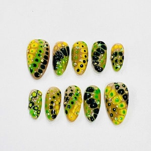 May include: Ten false nails with a green, black, and yellow abstract design. The nails are oval shaped and have a glossy finish.