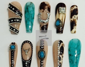 May include: A collection of ten coffin-shaped false nails with various designs. The nails feature a range of colors, including beige, turquoise, and brown, and are adorned with embellishments like rhinestones, a horseshoe, and a bull skull. The nails are arranged on a white surface.