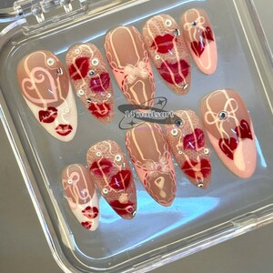 Pode incluir: Um conjunto de unhas posti&ccedil;as em uma caixa de pl&aacute;stico transparente. As unhas apresentam v&aacute;rios designs, incluindo l&aacute;bios vermelhos, cora&ccedil;&otilde;es e padr&otilde;es de renda. Algumas unhas t&ecirc;m uma base nude com detalhes vermelhos, enquanto outras s&atilde;o brancas com detalhes vermelhos. O texto "14 nailsart" &eacute; vis&iacute;vel.