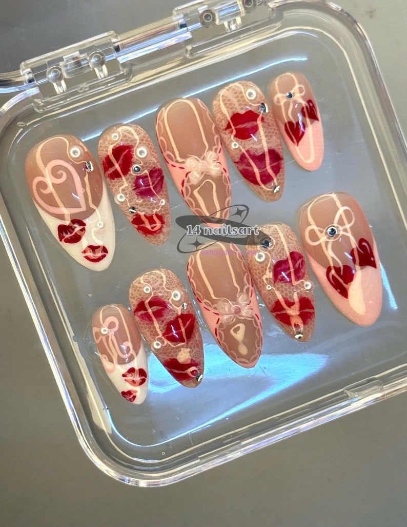 Pode incluir: Um conjunto de unhas posti&ccedil;as em uma caixa de pl&aacute;stico transparente. As unhas apresentam v&aacute;rios designs, incluindo l&aacute;bios vermelhos, cora&ccedil;&otilde;es e padr&otilde;es de renda. Algumas unhas t&ecirc;m uma base nude com detalhes vermelhos, enquanto outras s&atilde;o brancas com detalhes vermelhos. O texto "14 nailsart" &eacute; vis&iacute;vel.