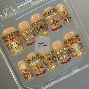 May include: A set of ten artificial nails in a clear plastic case. The nails are a nude colour and adorned with gold-coloured embellishments, including chains, jewels, and clock faces. The design is intricate and detailed.