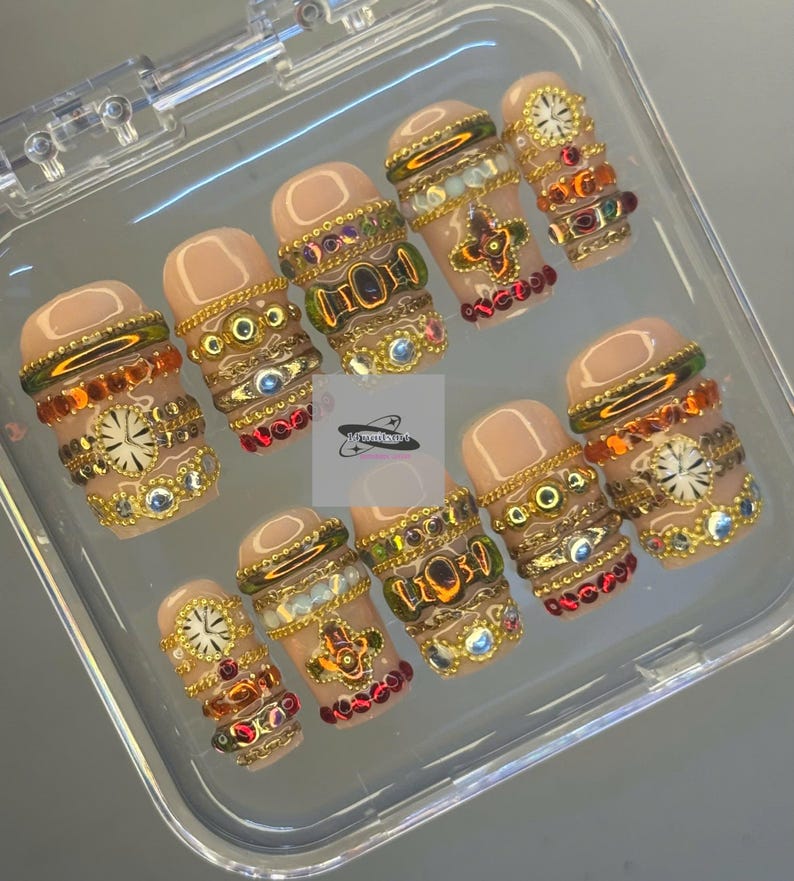 May include: A set of ten artificial nails in a clear plastic case. The nails are a nude colour and adorned with gold-coloured embellishments, including chains, jewels, and clock faces. The design is intricate and detailed.
