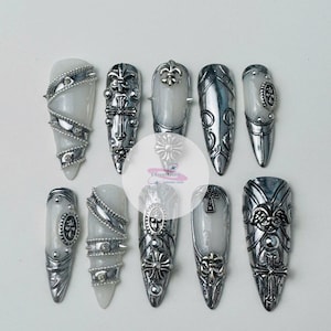 May include: A set of ten long, silver and white press-on nails. Each nail features intricate silver embellishments, including crosses, fleur-de-lis, and ornate designs. The nails have a glossy finish and a pointed shape.