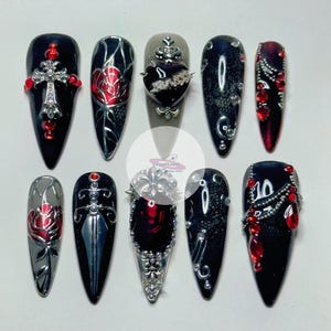 May include: A set of ten long, pointed, artificial nails. Each nail features a dark base color, with embellishments including crosses, roses, hearts, and ornate silver and red accents. The nails are designed with a gothic aesthetic.