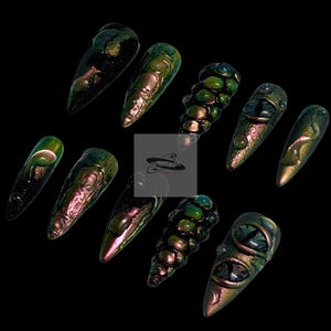Peut inclure: Dix faux ongles iris&eacute;s et pointus. Les ongles pr&eacute;sentent divers motifs, notamment des textures et des d&eacute;tails en forme d'yeux. La palette de couleurs oscille entre le vert, le violet et l'or, sur fond noir.