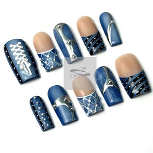 May include: A set of ten artificial nails with a denim-inspired design. The nails feature various embellishments, including silver-colored metal accents, white lace-like patterns, and black stripes. Some nails have a natural-toned base.
