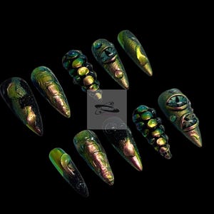 Peut inclure: Un ensemble de dix faux ongles longs et pointus. Les ongles sont principalement verts et dor&eacute;s avec des effets iris&eacute;s. Certains ongles pr&eacute;sentent un motif d'&oelig;il, tandis que d'autres ont des motifs textur&eacute;s en forme d'&eacute;cailles. Le fond est noir.