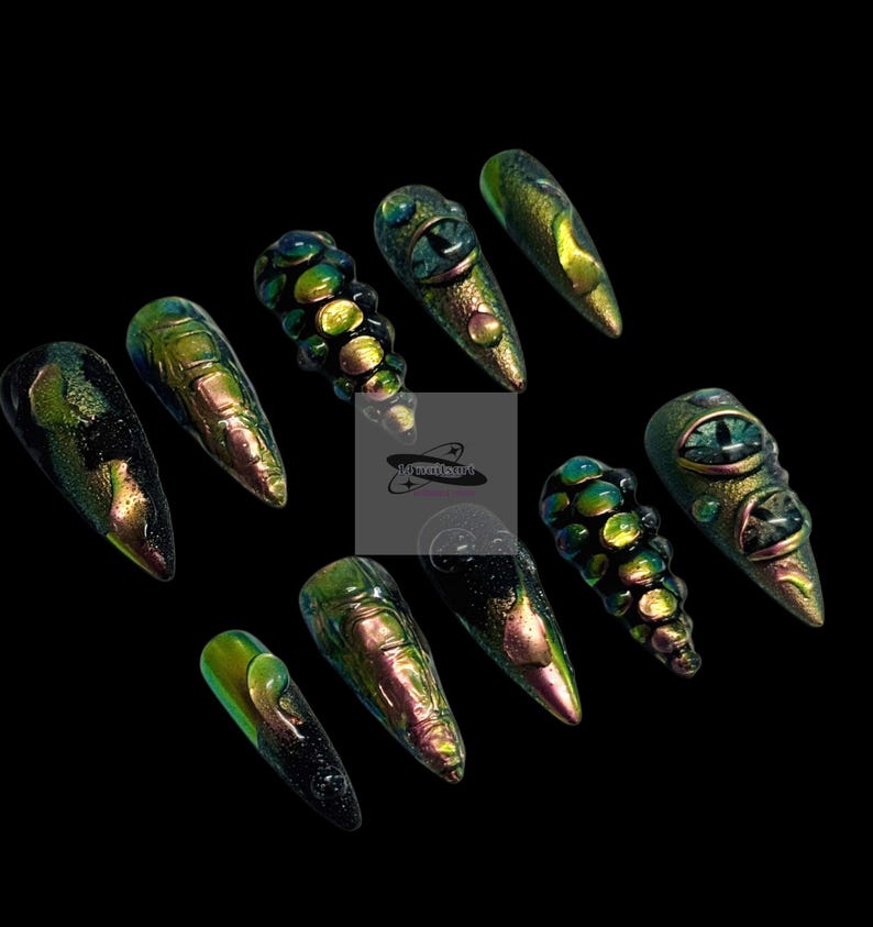 Peut inclure: Un ensemble de dix faux ongles longs et pointus. Les ongles sont principalement verts et dor&eacute;s avec des effets iris&eacute;s. Certains ongles pr&eacute;sentent un motif d'&oelig;il, tandis que d'autres ont des motifs textur&eacute;s en forme d'&eacute;cailles. Le fond est noir.