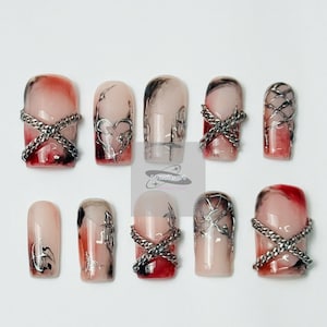 May include: A set of ten artificial nails with a variety of designs. Some nails feature a chain accent, while others have silver heart or abstract designs. The nails have a gradient effect with red and pink hues, and some have black accents.