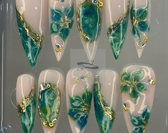 May include: A set of ten long, almond-shaped false nails. Each nail features a pale pink base with intricate designs in shades of teal and gold. Some nails have gold foil accents and small, sparkling rhinestones.
