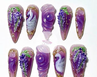 May include: A collection of ten long, pointed artificial nails. Each nail showcases a distinct design, such as purple wisteria, a white swan, and swirling purple patterns. Gold trim and tiny pearls add to the elegant, artistic aesthetic of the nail art.
