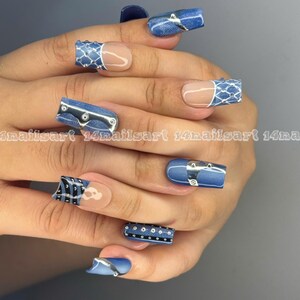 May include: A hand with long, square-shaped acrylic nails. The nails feature a denim-inspired design with silver accents, including rivets and belt-like details. Some nails have a clear base with a white and blue pattern.