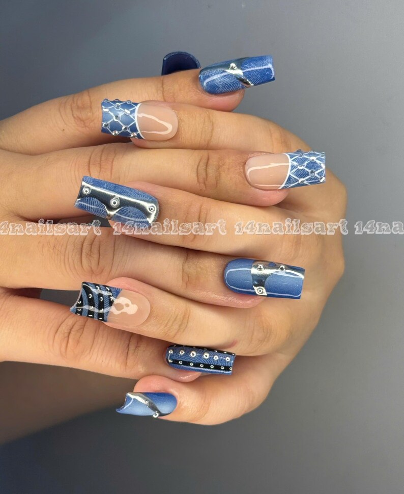 May include: A hand with long, square-shaped acrylic nails. The nails feature a denim-inspired design with silver accents, including rivets and belt-like details. Some nails have a clear base with a white and blue pattern.