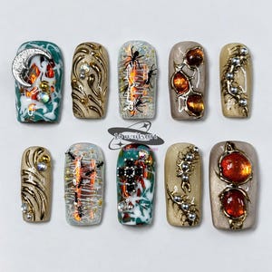 May include: Ten press-on nails featuring intricate designs including a silver crescent moon, gold swirls, black ant silhouettes, amber-colored gems, and metallic beads. The set includes the logo 14nailsart handmade luxury in the center.