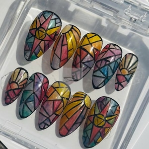 May include: A set of ten long, oval-shaped false nails. Each nail is painted with a stained glass design featuring a sunburst or geometric patterns in shades of yellow, red, blue, and purple, outlined in black.