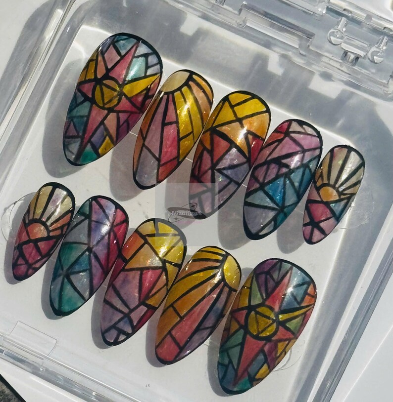 May include: A set of ten long, oval-shaped false nails. Each nail is painted with a stained glass design featuring a sunburst or geometric patterns in shades of yellow, red, blue, and purple, outlined in black.