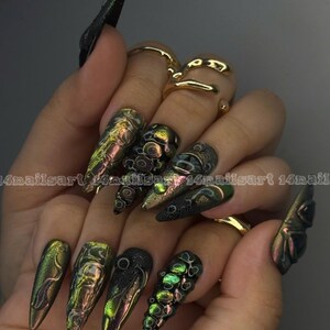Peut inclure: Une main pr&eacute;sente une manucure longue et pointue, aux couleurs iris&eacute;es vertes, dor&eacute;es et noires. Certains ongles arborent des motifs textur&eacute;s et des embellissements en 3D. Des bagues dor&eacute;es ornent les doigts. Le texte "14nailsart 14nail" est visible.