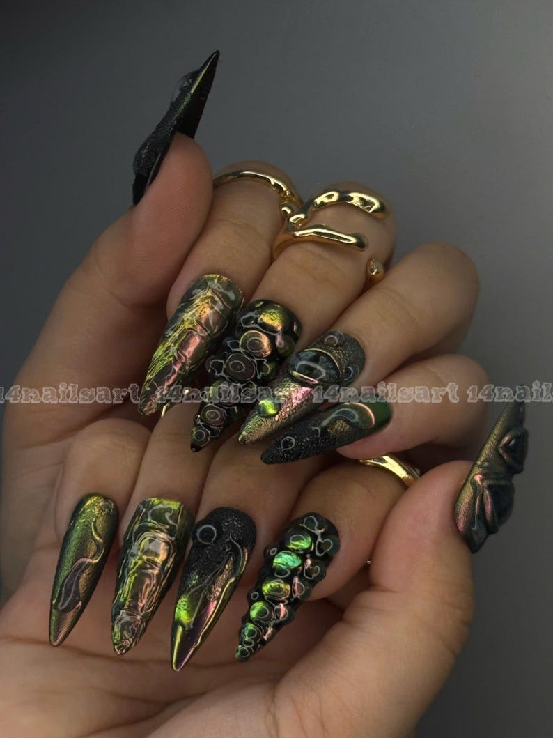 Peut inclure: Une main pr&eacute;sente une manucure longue et pointue, aux couleurs iris&eacute;es vertes, dor&eacute;es et noires. Certains ongles arborent des motifs textur&eacute;s et des embellissements en 3D. Des bagues dor&eacute;es ornent les doigts. Le texte "14nailsart 14nail" est visible.