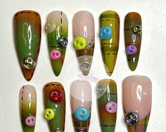 May include: Ten long, almond-shaped false nails with a green, brown, and plaid design. Each nail has a different colored button on top.