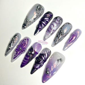 May include: Set of ten long, pointed, iridescent purple and silver press-on nails with various designs, including swirls, flowers, and abstract patterns.