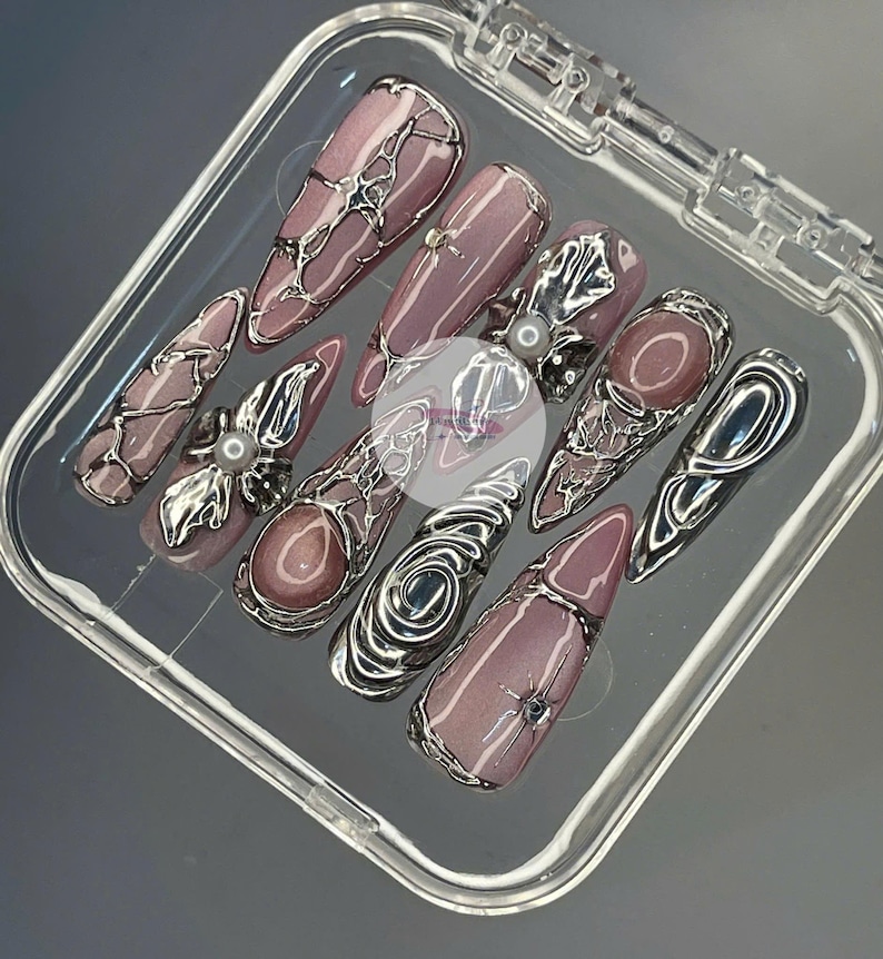 May include: A set of ten artificial nails in a clear plastic case. The nails are a mauve color with silver metallic embellishments. Some nails have a pearl accent. The nails are long and pointed.
