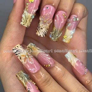 May include: A hand displaying long, decorated fingernails. Each nail features a pink base with intricate designs, including gold filigree, seashells, and floral accents. Some nails have a zebra print pattern. The text "14nailart" is visible.