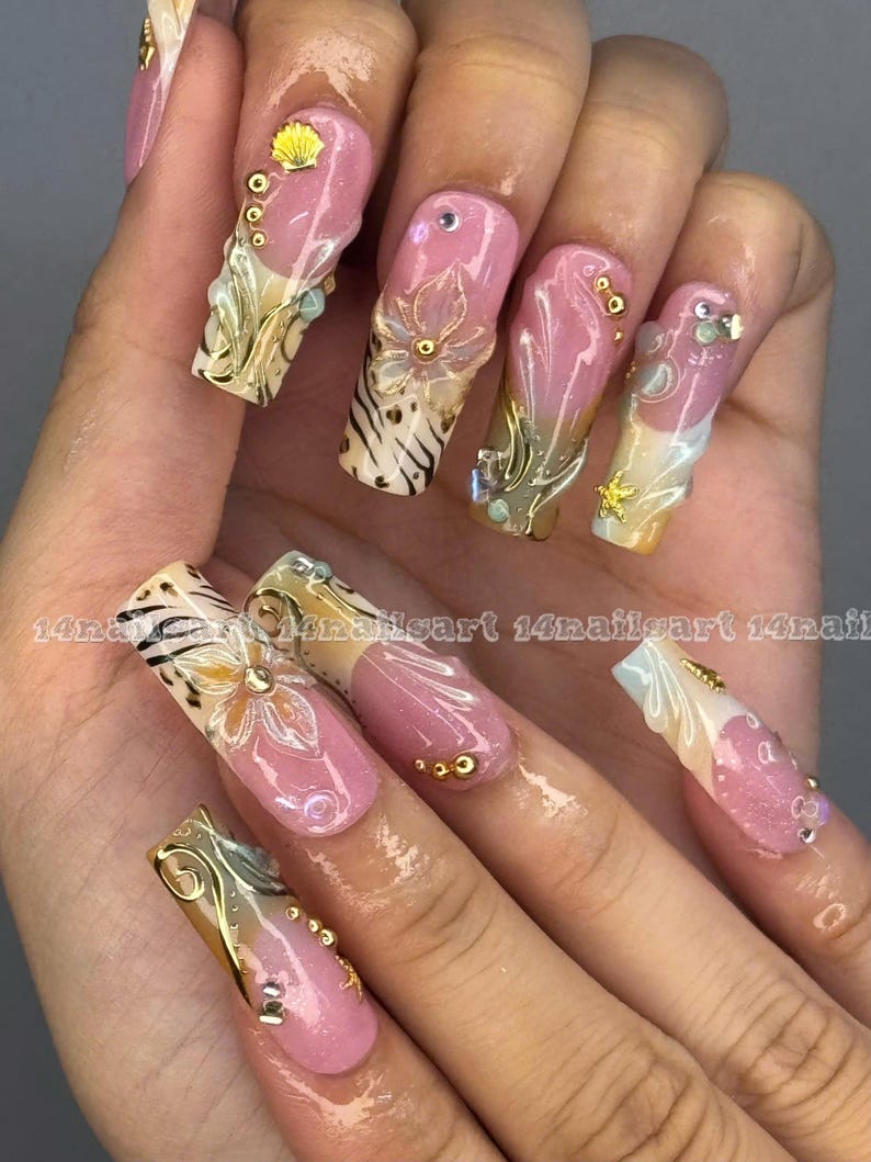 May include: A hand displaying long, decorated fingernails. Each nail features a pink base with intricate designs, including gold filigree, seashells, and floral accents. Some nails have a zebra print pattern. The text "14nailart" is visible.