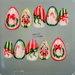 May include: A set of ten artificial nails with Christmas-themed designs. The nails feature images of wreaths, bows, candy canes, and a nutcracker soldier. The color palette includes red, green, white, and gold accents, creating a festive look.
