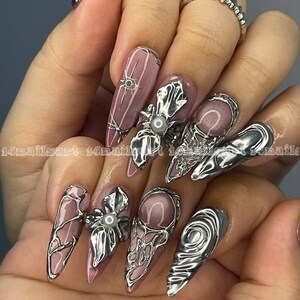 May include: A hand displaying long, almond-shaped nails with a mauve base color. Each nail features intricate silver embellishments, including floral designs, geometric patterns, and pearl accents. The nails are adorned with the text "14nailsart".