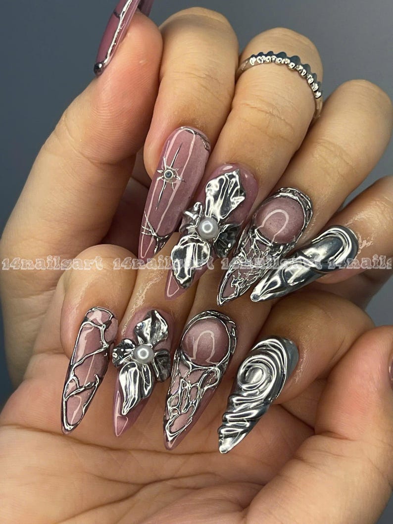 May include: A hand displaying long, almond-shaped nails with a mauve base color. Each nail features intricate silver embellishments, including floral designs, geometric patterns, and pearl accents. The nails are adorned with the text "14nailsart".