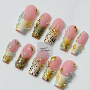 May include: A set of ten artificial nails with a pink and white gradient design. Each nail features intricate embellishments, including gold accents, small beads, and floral or seashell-inspired decorations. The nails have a glossy finish.